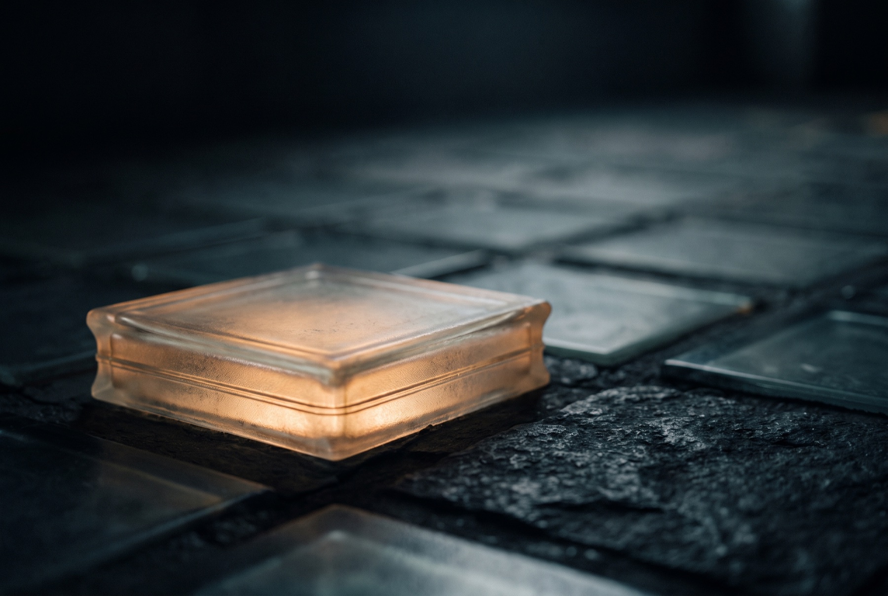 One raised frosted glass tile in close-up, edge-lit, thin bands of warm light stacked inside it, the flat tiles dark behind.